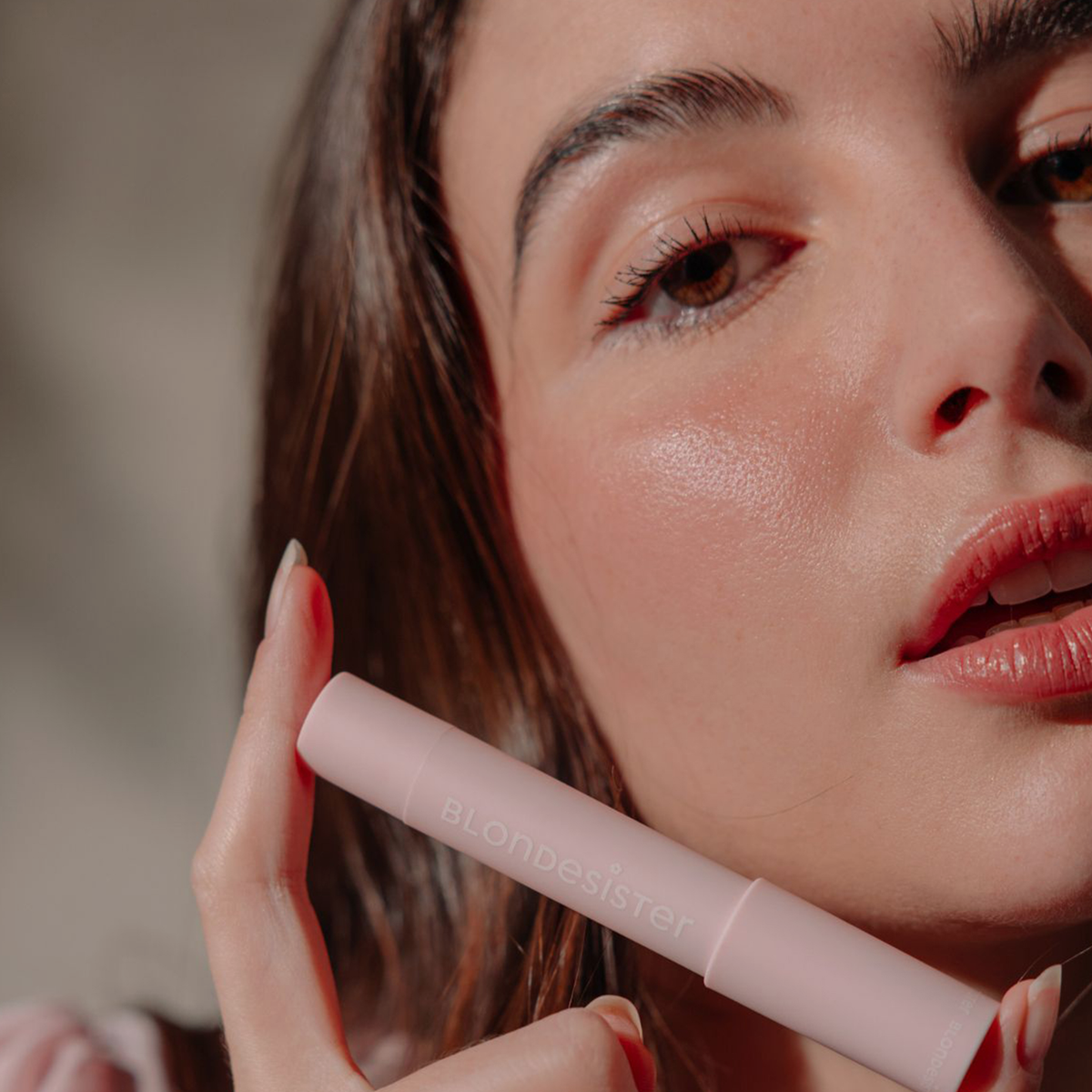 Eine Frau mit strahlender Haut hält eine rosa Tube „BLONDESISTER 2 IN 1 IT’S UP TO YOU LIP OR CHEEK“ in sanftem, natürlichem Licht nahe an ihr Gesicht.
