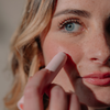 Eine Person trägt den „BLONDESISTER 2 IN 1 IT’S UP TO YOU LIP OR CHEEK“-Stift auf ihrer Wange auf und bringt so ihre klare Haut und ihre blauen Augen zur Geltung.