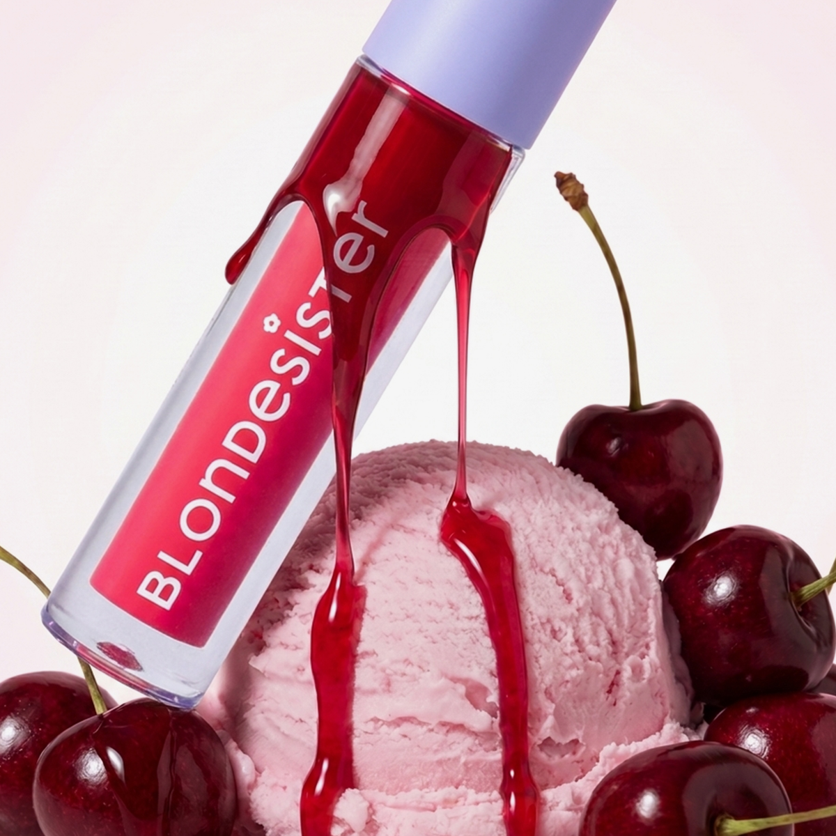 BLONDESISTER 2-IN-1 SWEET GLOSS GETÖNTES LIPPENÖL tropft auf rosa Eis, umgeben von Kirschen.