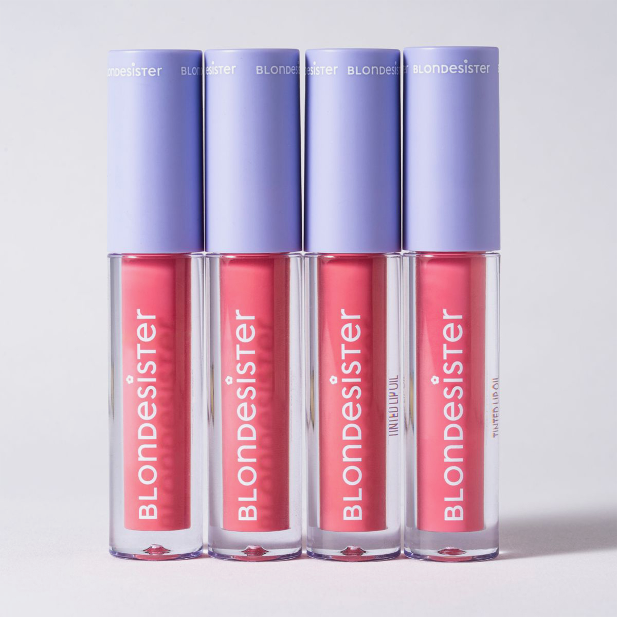 Vier Tuben „BLONDESISTER 2 IN 1 SWEET GLOSS TINTED LIP OIL“ mit rosa Glanz und violetten Verschlusskappen sind vor einem hellen Hintergrund zu sehen.