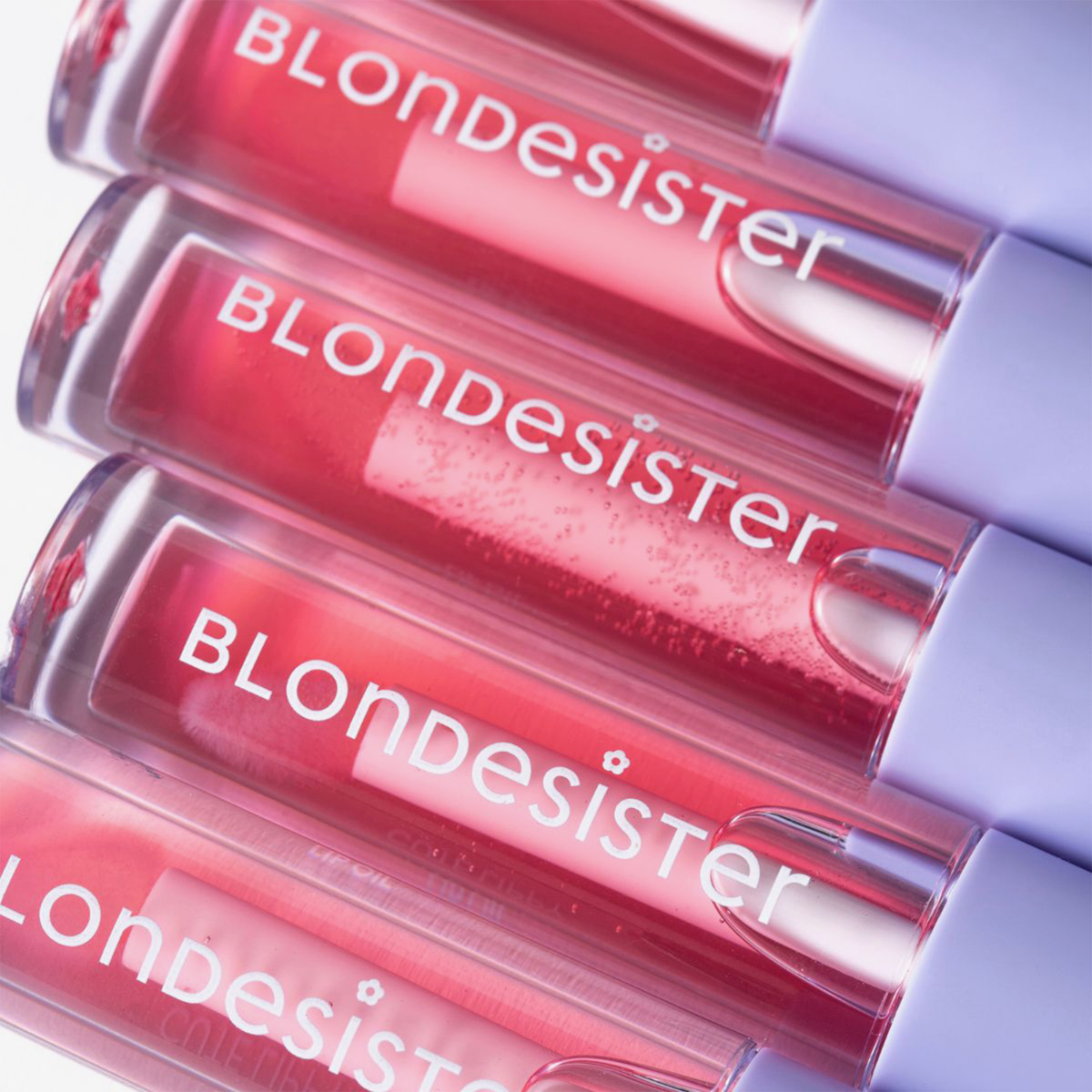Fünf Tuben „BLONDESISTER 3 IN 1 CUTE LIPPY LIP OIL“ mit weißen Verschlusskappen sind diagonal angeordnet, aus der Nahaufnahme betrachtet.