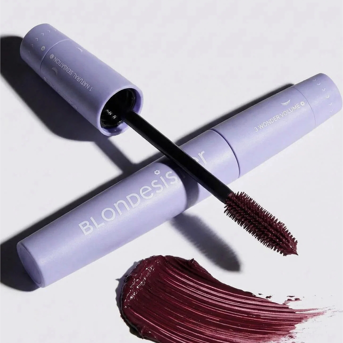 Zwei Tuben „BLONDESISTER 4 IN 1 CLICK & TURN MASCARA“, von denen eine geöffnet ist und dunkle, burgunderrote Wimperntusche sowie einen Schmierfleck darunter zeigt.