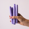 Eine Hand mit rot lackierten Fingernägeln hält vier violette BLONDESISTER 4 IN 1 CLICK & TURN MASCARA-Tuben vor einem einfarbigen Hintergrund.