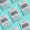 Mehrere weiße und blaue rechteckige Verpackungen mit Emergency Dots-Pflastern mit Teebaumöl zur Abdeckung von Hautunreinheiten und Pickeln auf hellblauem Hintergrund.