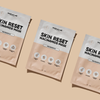 Drei rechteckige Breakout+Aid Skin Reset Niacinamide Mask-Päckchen, die diagonal auf einem hellbraunen Untergrund angeordnet sind.