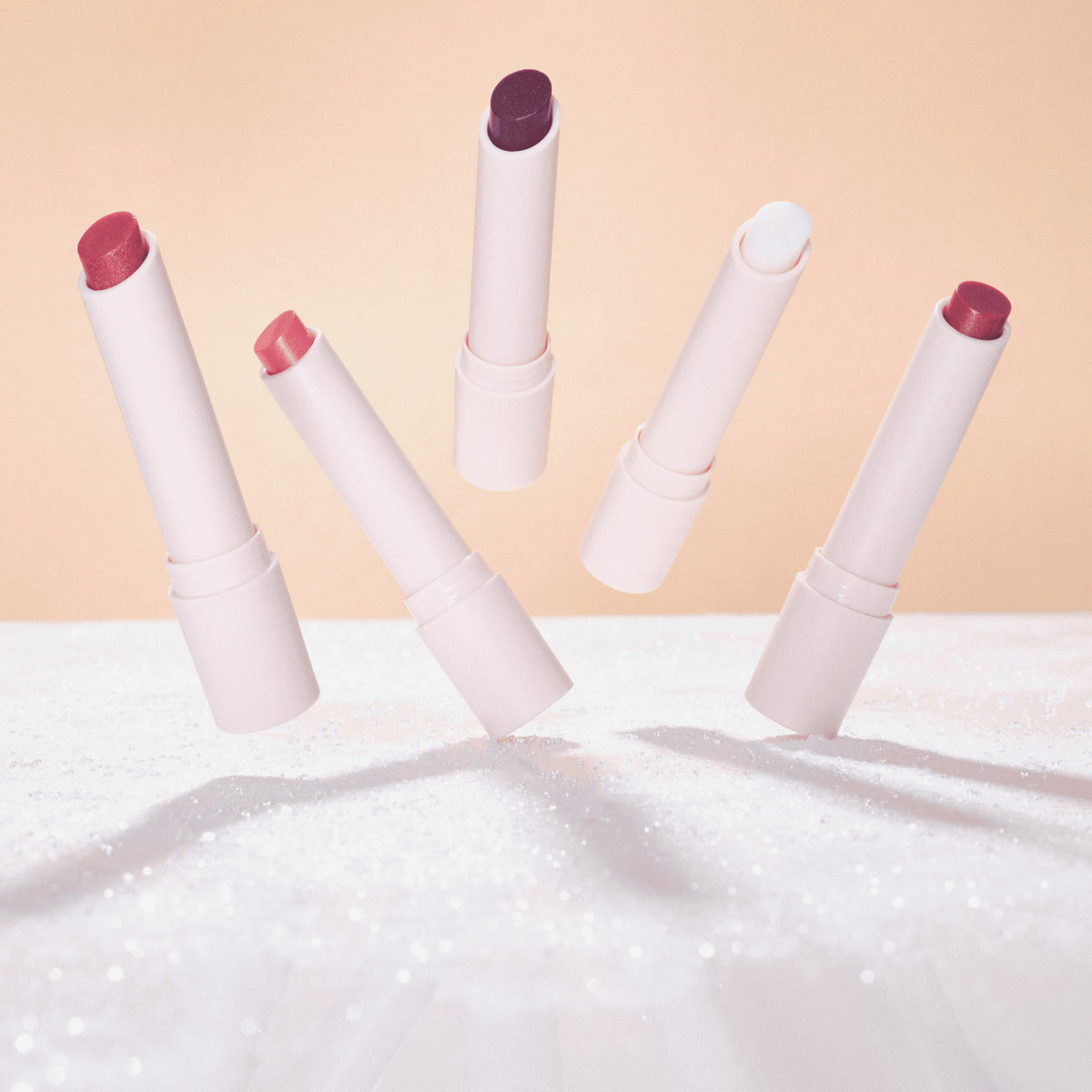 Fünf schimmernde Catrice Diamond Glaze Gloss Sticks in verschiedenen Farben, scheinbar schwebend, vor einem warmen Hintergrund auf glitzernder Fläche.