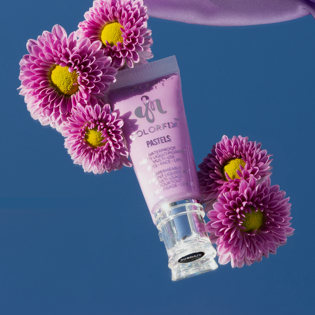Eine Tube Danessa Myricks Beauty Colorfix Pastels Sundaze in pastelligem Lila ist von rosa Gänseblümchen auf blauem Hintergrund umgeben.