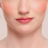 Eine Nahaufnahme von glänzenden rosa Lippen und pfirsichfarbenem Rouge mit dem Juicy Lip Balm von Dominique Cosmetics.