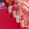 Vier Tuben Essence GLOSSY GLAZE High Shine Lipstick in verschiedenen Farbtönen sind auf roten Stufen mit einem lebendigen, bunten Hintergrund angeordnet.