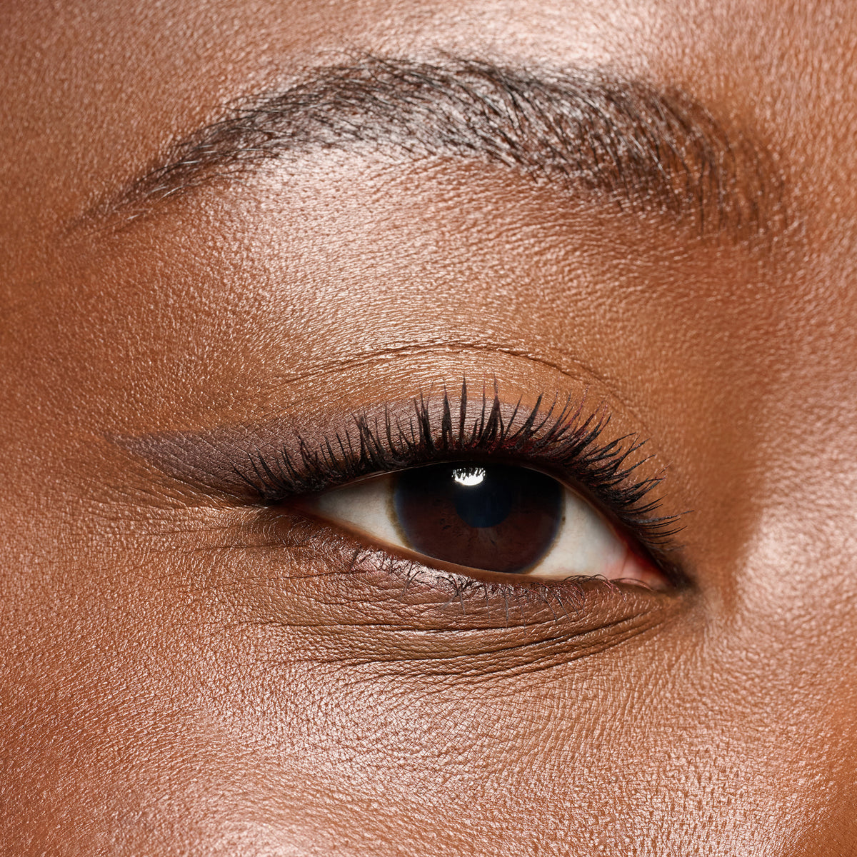 Nahaufnahme eines braunen Auges mit sanftem braunem Eyeliner und langen, geschwungenen Wimpern auf glatter Haut.