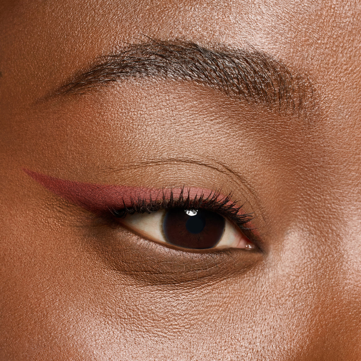 Nahaufnahme eines braunen Auges mit sanftem burgunderfarbenem Eyeliner und langen, geschwungenen Wimpern auf glatter Haut.