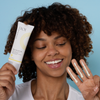 Eine lächelnde Frau hält Geek & Gorgeous Zero Feel SPF 50 in der Hand und zeigt Farbmuster auf ihren Fingern vor einem blauen Hintergrund.