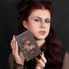 Eine Person mit langen, dunkelroten Haaren hält die Palette „Over The Garden Wall – Tome of the Unknown“ von Lethal Cosmetics vor einem schwarzen Hintergrund.