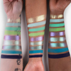 Auf drei Unterarmen sind Farbmuster der „The Legend of Korra – Raava Palette“ von Lethal Cosmetics in Pastell- und Metallic-Streifen zu sehen.