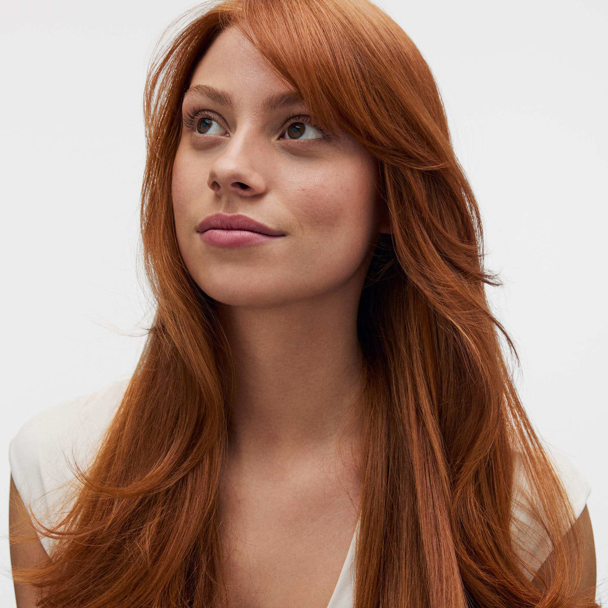 Eine Frau mit langen roten Haaren und heller Haut blickt nach oben. Sie trägt ein weißes Oberteil vor einem unifarbenen Hintergrund.