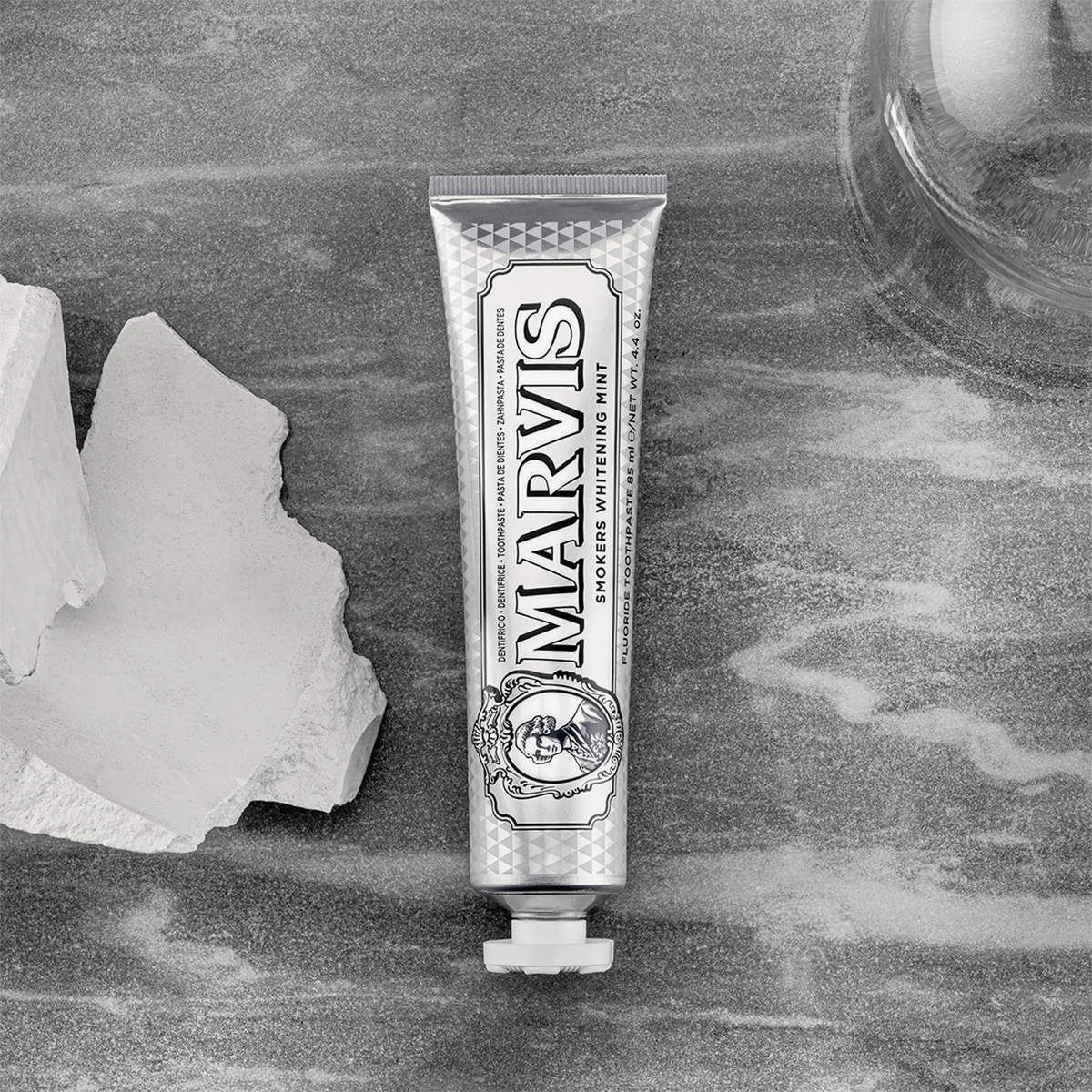 Eine Tube Marvis Zahnpasta Smokers Whitening Mint liegt auf einer grauen Fläche neben weißen Steinen und einem Glasobjekt.