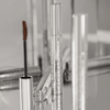 Femuline™ Brownie Mascara von Mesauda Beauty mit Zauberstab, gespiegelt auf einer glänzenden Oberfläche mit verschwommenen Tuben im Hintergrund.