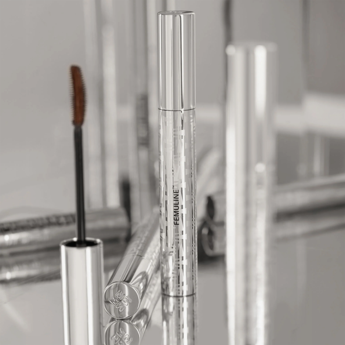 Femuline™ Brownie Mascara von Mesauda Beauty mit Zauberstab, gespiegelt auf einer glänzenden Oberfläche mit verschwommenen Tuben im Hintergrund.