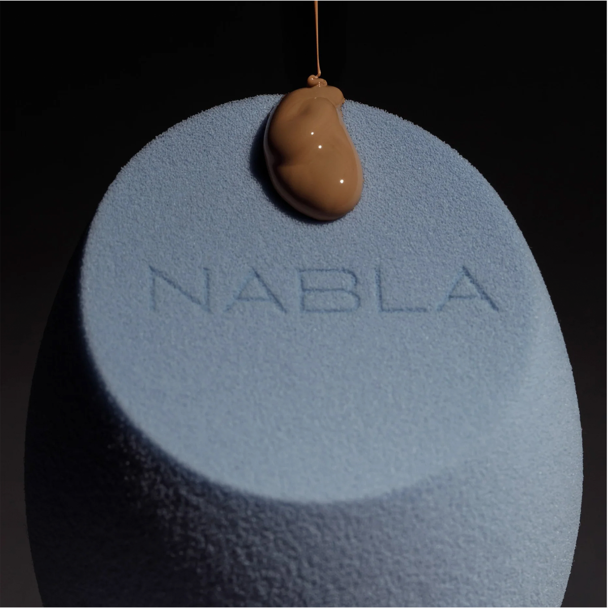 Der Blended-up Pro Tapered Sponge von NABLA Cosmetics, blau mit geprägtem Logo, ist mit flüssiger Foundation bestückt.