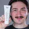 Lächelnder Mann mit einer Tube der Nø Cosmetics basic tøday Base Hydration Cream in der Hand und einem Tupfer auf der Wange.