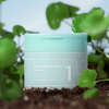 Ein Tiegel numbuzin No.1 Centella Re-Leaf Green Toner Pad ruht auf dem Boden, umgeben von üppigen grünen Blättern und Pflanzen.