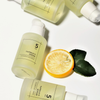 Vier Flaschen des numbuzin No.5 Goodbye Blemish Serum sind mit einer Zitronenscheibe und zwei grünen Blättern auf weißem Hintergrund abgebildet.