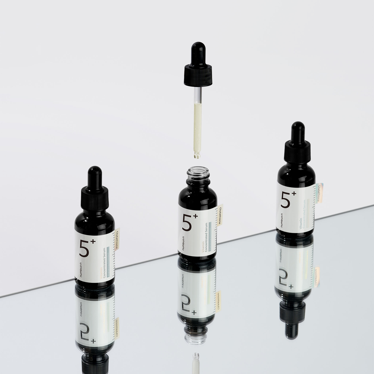 Abgebildet sind drei schwarze Fläschchen mit No.5+ Vitamin Concentrated Serum, wobei ein Tropfer über einem offenen Fläschchen auf einer verspiegelten Fläche gehalten wird.