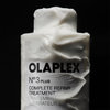 Eine Flasche OLAPLEX N°3 PLUS Complete Repair Treatment, umhüllt von einer dicken weißen Creme, auf schwarzem Hintergrund.