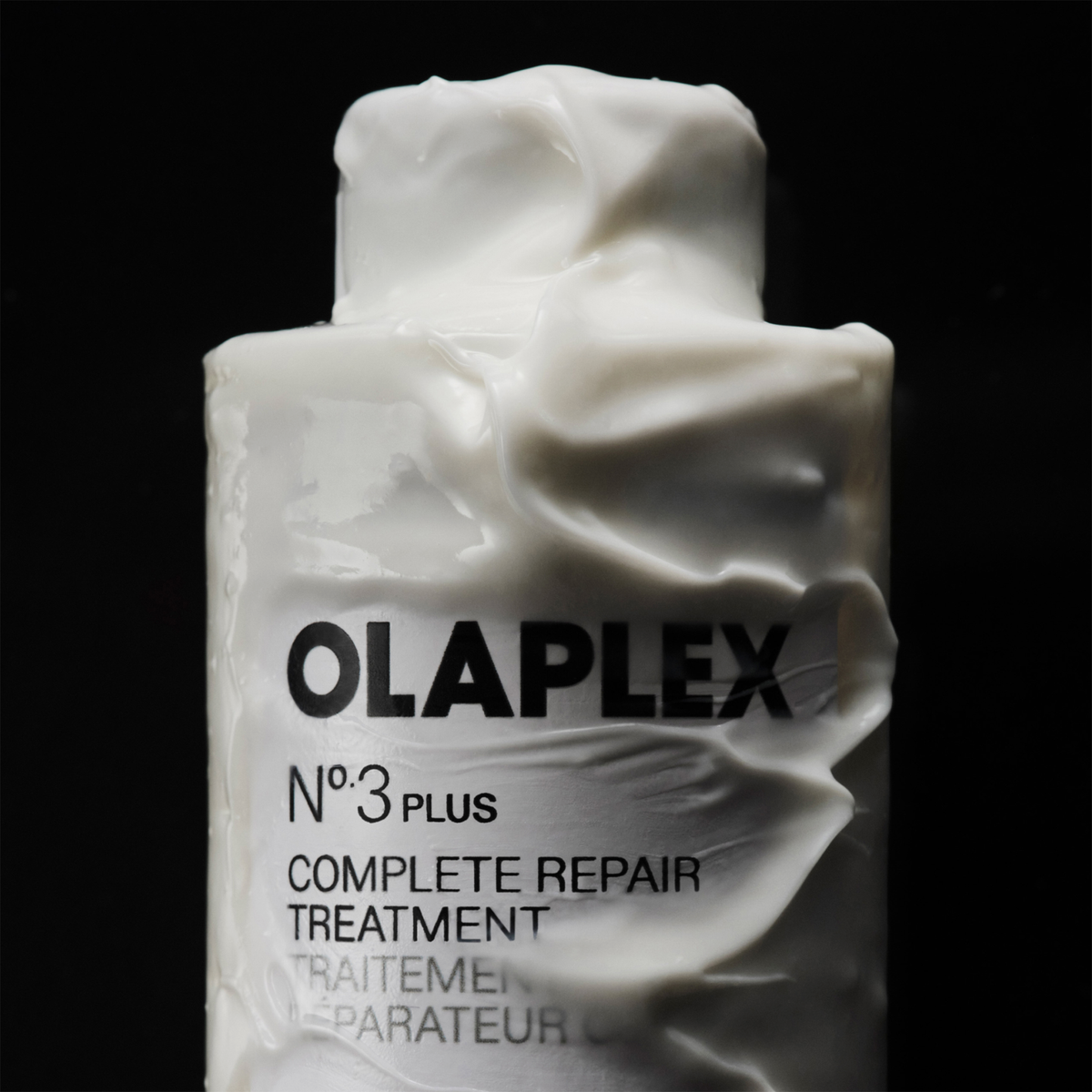 Eine Flasche OLAPLEX N°3 PLUS Complete Repair Treatment, umhüllt von einer dicken weißen Creme, auf schwarzem Hintergrund.