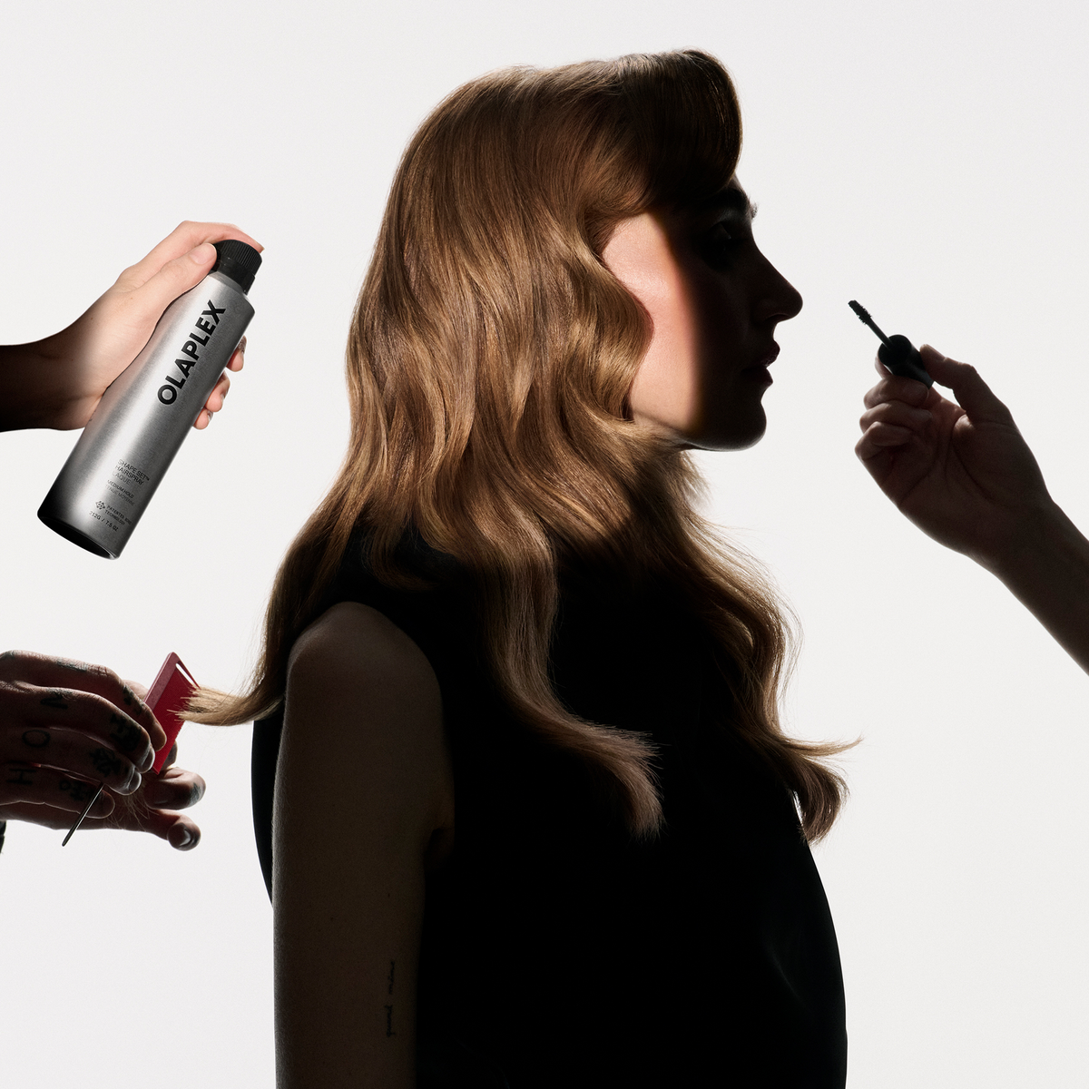 Eine Frau mit gewelltem Haar, deren Frisur von drei Händen gestylt wird, die eine Flasche OLAPLEX Shape Set Hairspray, einen roten Kamm und einen Mascara-Bürstchen halten.