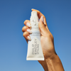 Eine Hand hält eine Tube „Philip Kingsley Finishing Touch Frizz Fighting Gloss“ vor einem strahlend blauen Himmel.
