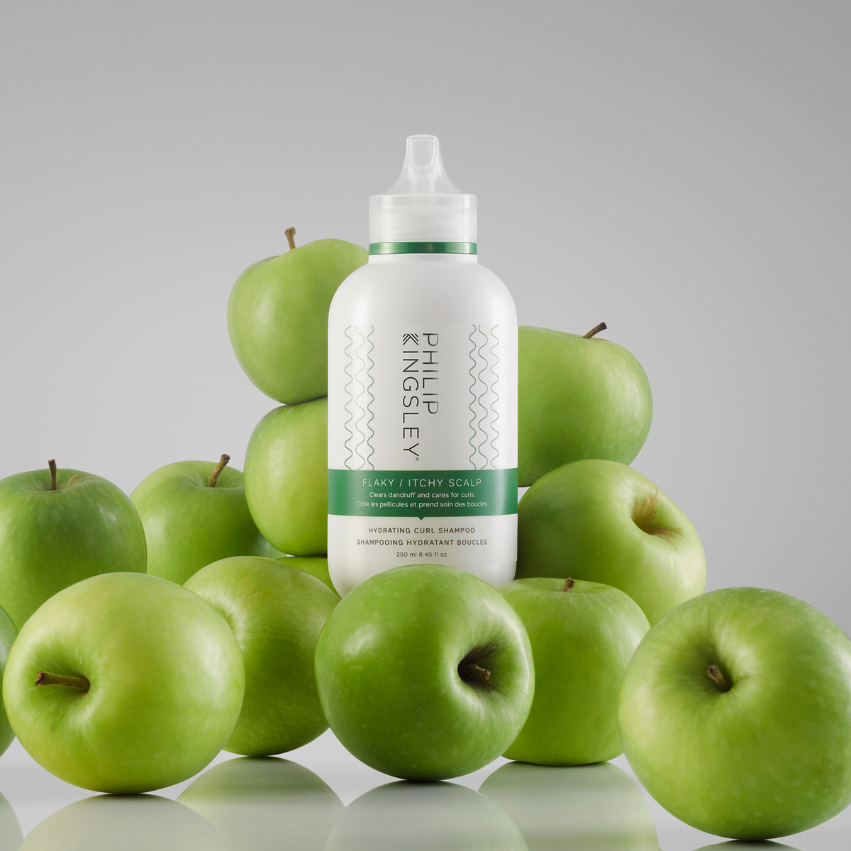 Eine Flasche „Philip Kingsley Flaky Itchy Scalp Shampoo“ steht auf einer spiegelnden Oberfläche, umgeben von grünen Äpfeln.