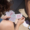 Eine Person hält im Freien Spielkarten, mit einer anderen Hand und Karten auf dem Boden, neben der PURISH Summer Bag 2025 – Deluxe Routine.