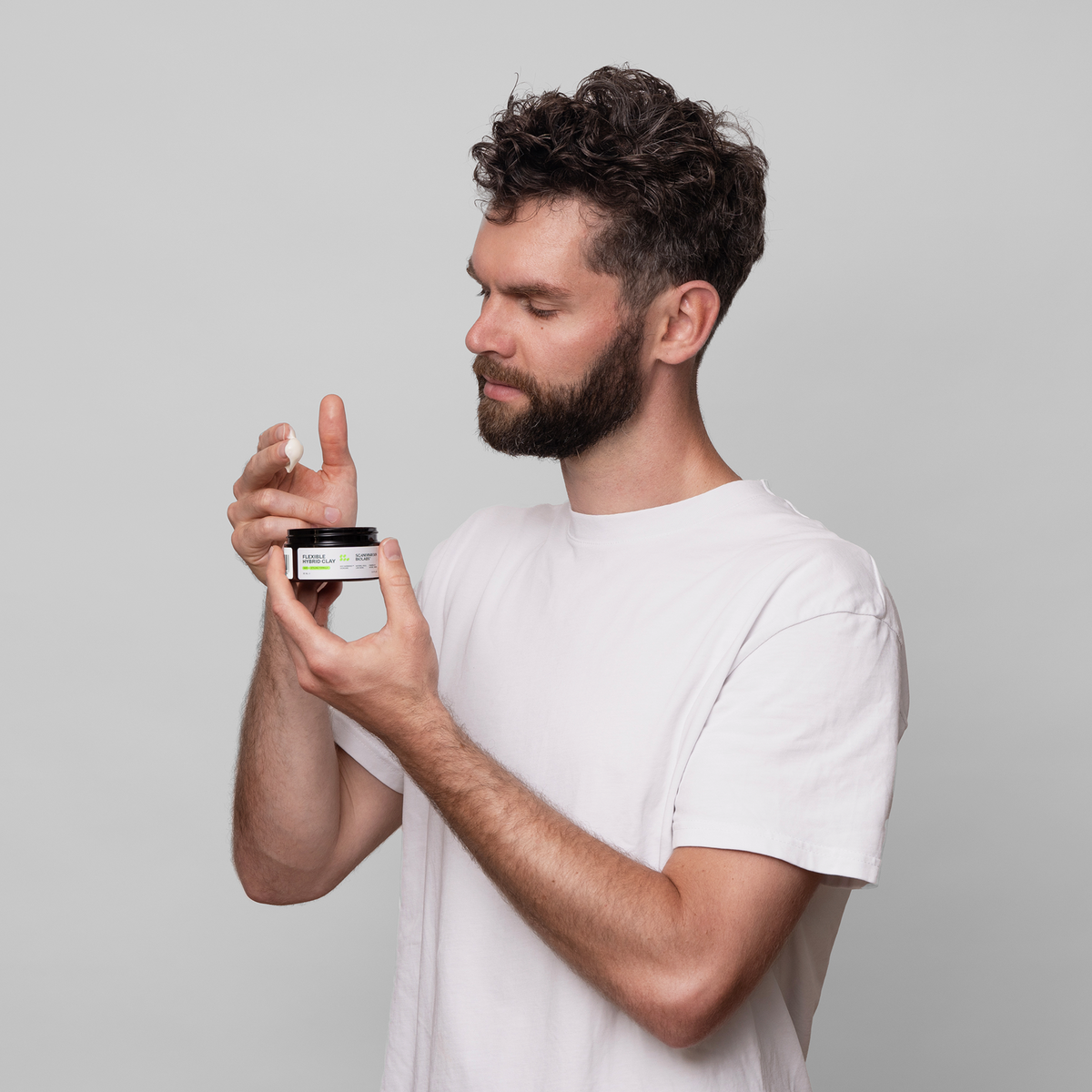 Ein Mann in einem weißen T-Shirt hält und öffnet ein Glas Scandinavian Biolabs Flexible Hybrid Clay und betrachtet es vor einem schlichten hellen Hintergrund.