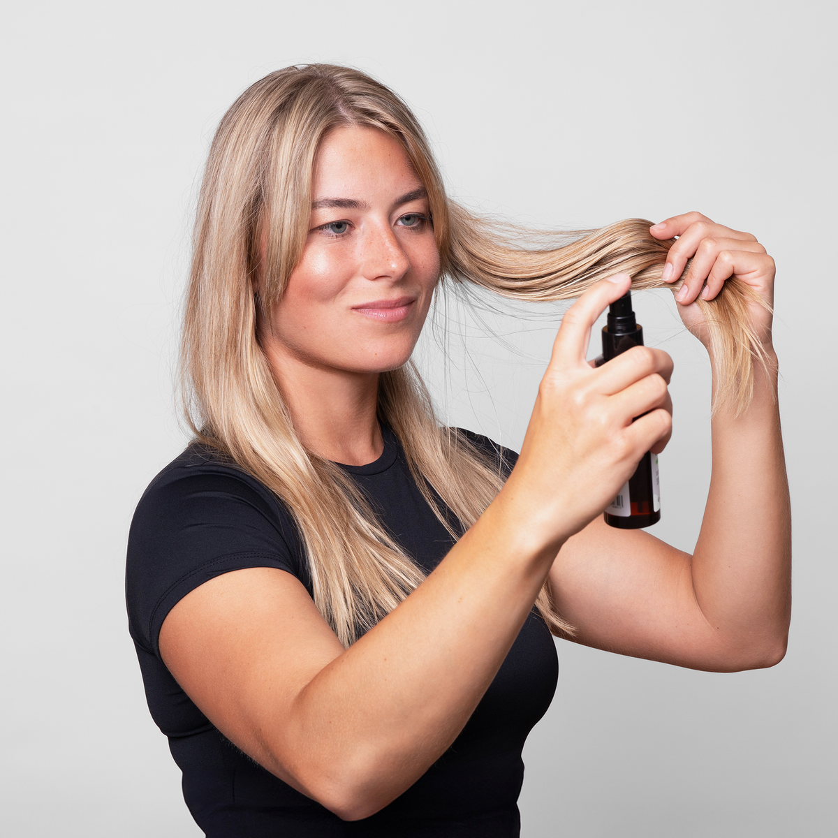 Eine Frau mit langem blondem Haar sprüht sich Scandinavian Biolabs Sea Mineral Spray auf ihr Haar und hält eine schwarze Flasche vor einem schlichten Hintergrund.