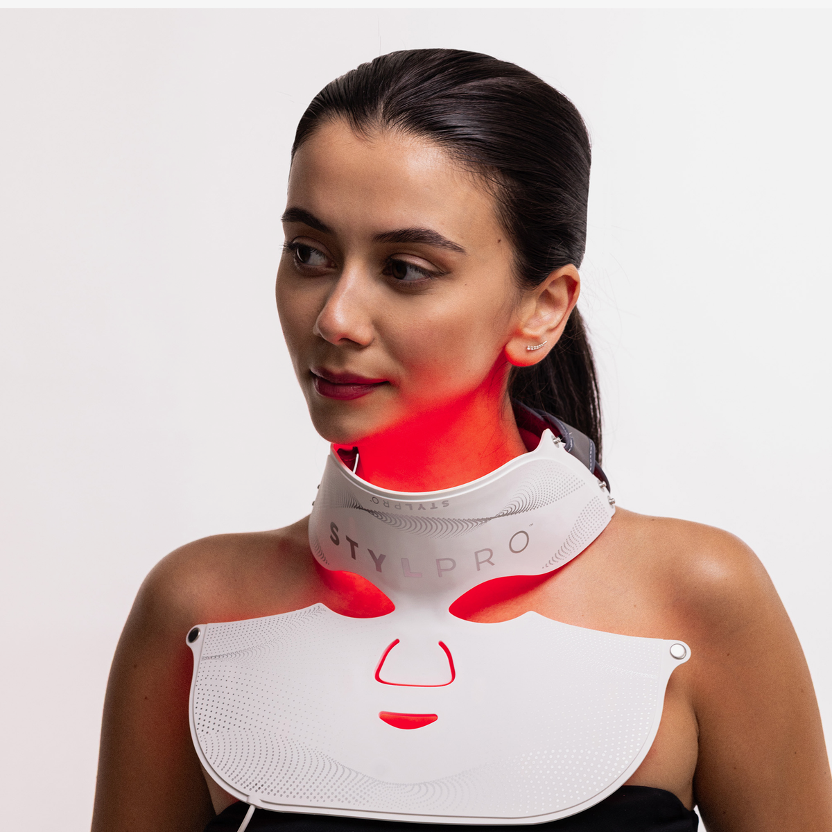 Eine Frau trägt die STYLPRO Wavelength Pro 5 in 1 LED-Maske, deren rote Lichter vor einem schlichten weißen Hintergrund leuchten um ihren Hals.