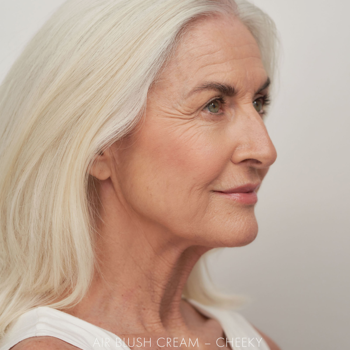 Eine reife Frau mit langen weißen Haaren trägt SWEED Air Blush Cream und lächelt sanft im Profil vor einem schlichten Hintergrund.