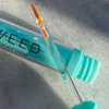 Eine türkisfarbene Tube des SWEED-Wimpernwachstumsserums mit Applikatorpinsel steht im Sonnenlicht auf einer hellgrauen Unterlage.