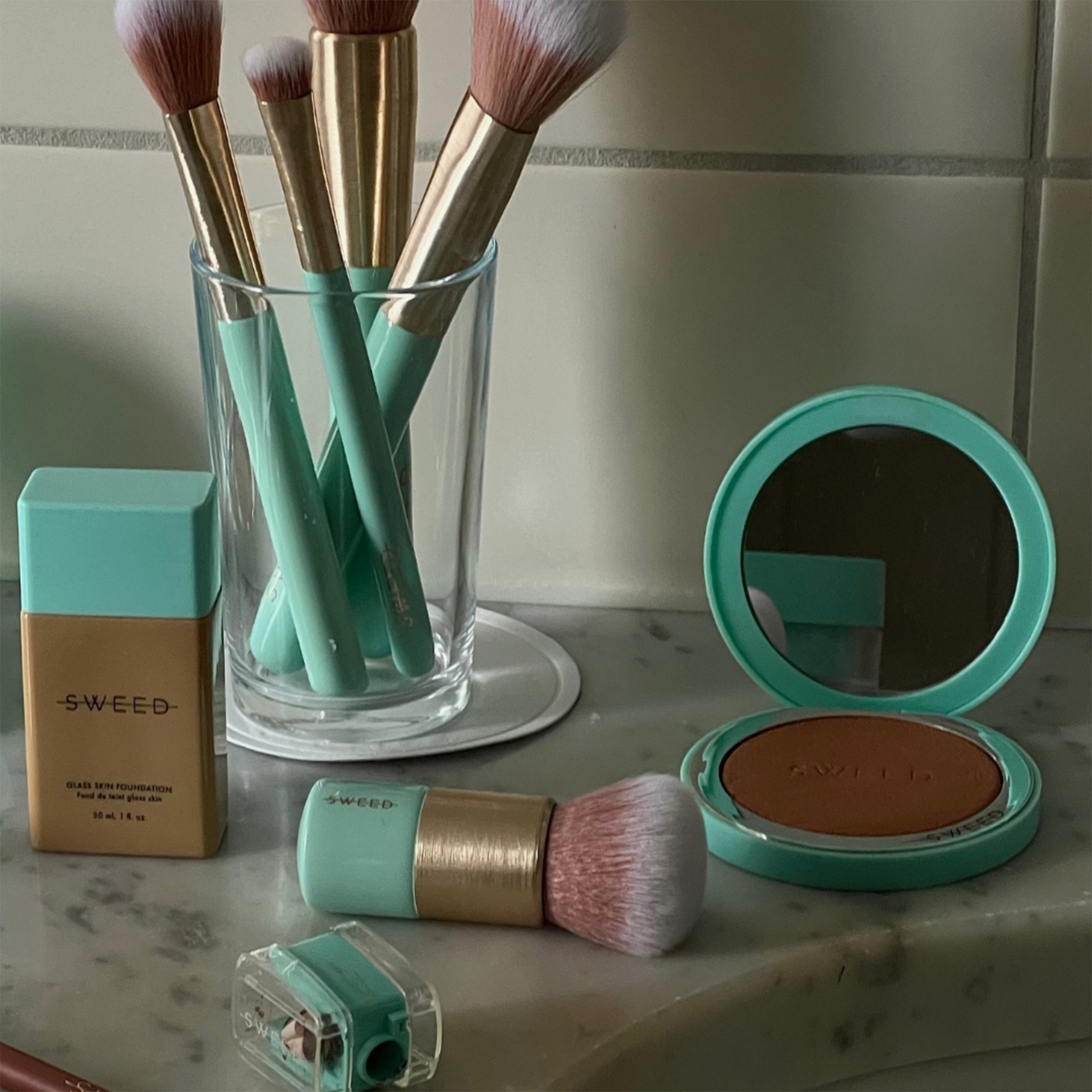 SWEED-Foundation-Pinsel, Puder-Kompakt und Anspitzer auf einer Marmorplatte zusammen mit anderen Make-up-Utensilien.