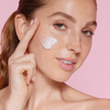 Vor einem rosa Hintergrund trägt eine lächelnde Frau mit zwei Fingern die Creme „Geek & Gorgeous Mighty Melt 150 ml“ auf ihre Wange auf.