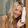 Eine lächelnde Frau mit langen blonden Haaren hält eine Flasche Bali Body Clear 1 Hour Express Tan neben ihr Gesicht.