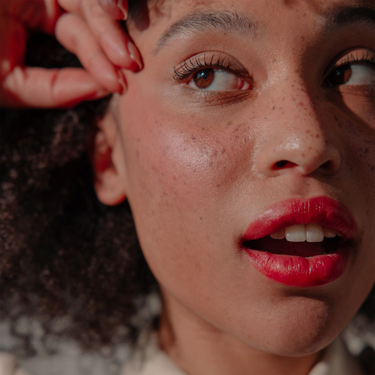Eine Nahaufnahme einer Person mit lockigem Haar, Sommersprossen und roten Lippen, die das Produkt „BLONDESISTER 2 IN 1 IT’S UP TO YOU Lip or Cheek“ verwendet.