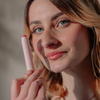Eine Frau mit hellen Augen lächelt und hält dabei „BLONDESISTER 2 IN 1 IT’S UP TO YOU LIP OR CHEEK“ in der Hand. Das Sonnenlicht lässt ihr Gesicht und ihr natürliches Make-up strahlen.