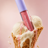 Eine Tube BLONDESISTER 2 IN 1 SWEET GLOSS TINTED LIP OIL zwischen zwei Kugeln schmelzendem Vanilleeis in einer Waffeltüte, vor einem rosa Hintergrund.