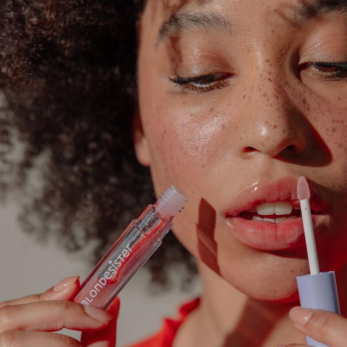 Eine Person mit lockigem Haar trägt das BLONDESISTER 3 IN 1 CUTE LIPPY LIP OIL mit einem Applikator auf, wobei sie die Tube nahe an ihre Lippen hält.