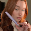 Eine Frau mit langen roten Haaren hält eine lilafarbene Tube „BLONDESISTER 4 IN 1 CLICK & TURN MASCARA“ in natürlichem Licht nahe an ihr Gesicht.