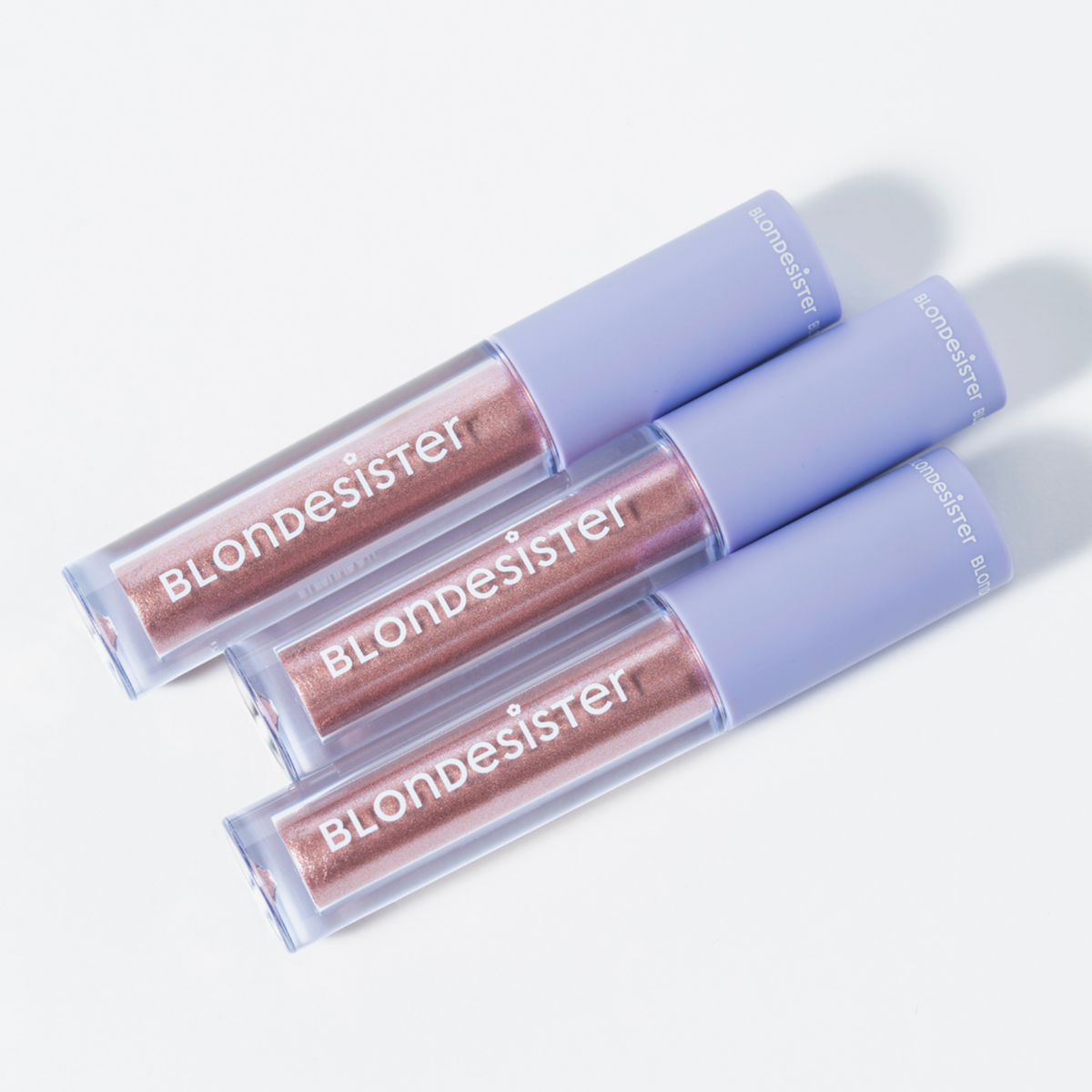 Drei BLONDESISTER GLOWEYE-Lidschattenstifte mit metallisch-roségoldenen und lavendelfarbenen Verschlusskappen stehen auf einer weißen Fläche in einer Reihe.