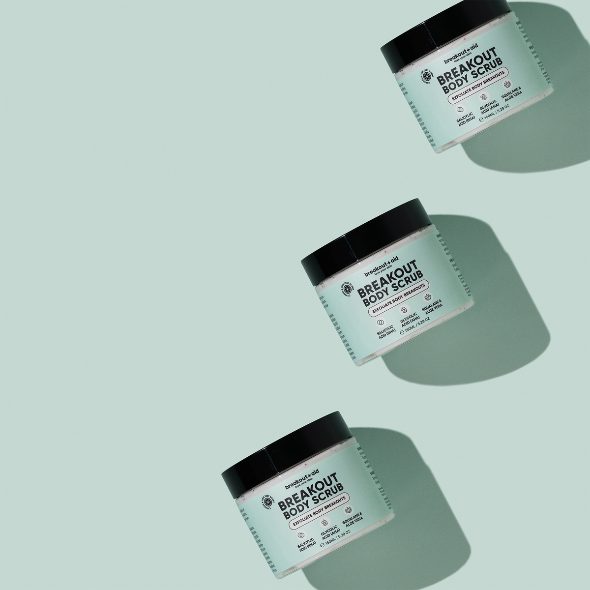 Drei Tiegel Breakout Body Scrub, diagonal auf einem hellgrünen Hintergrund angeordnet.