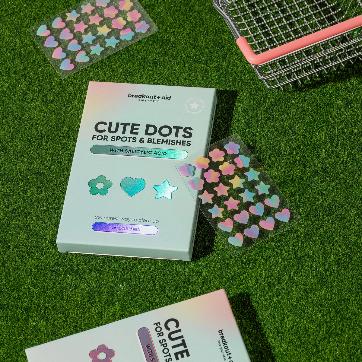 Breakout+aid Cute Dots – Akne-Pflaster gegen Pickel und Hautunreinheiten mit schillernden Stern- und Herz-Aufklebern auf grünem Gras.