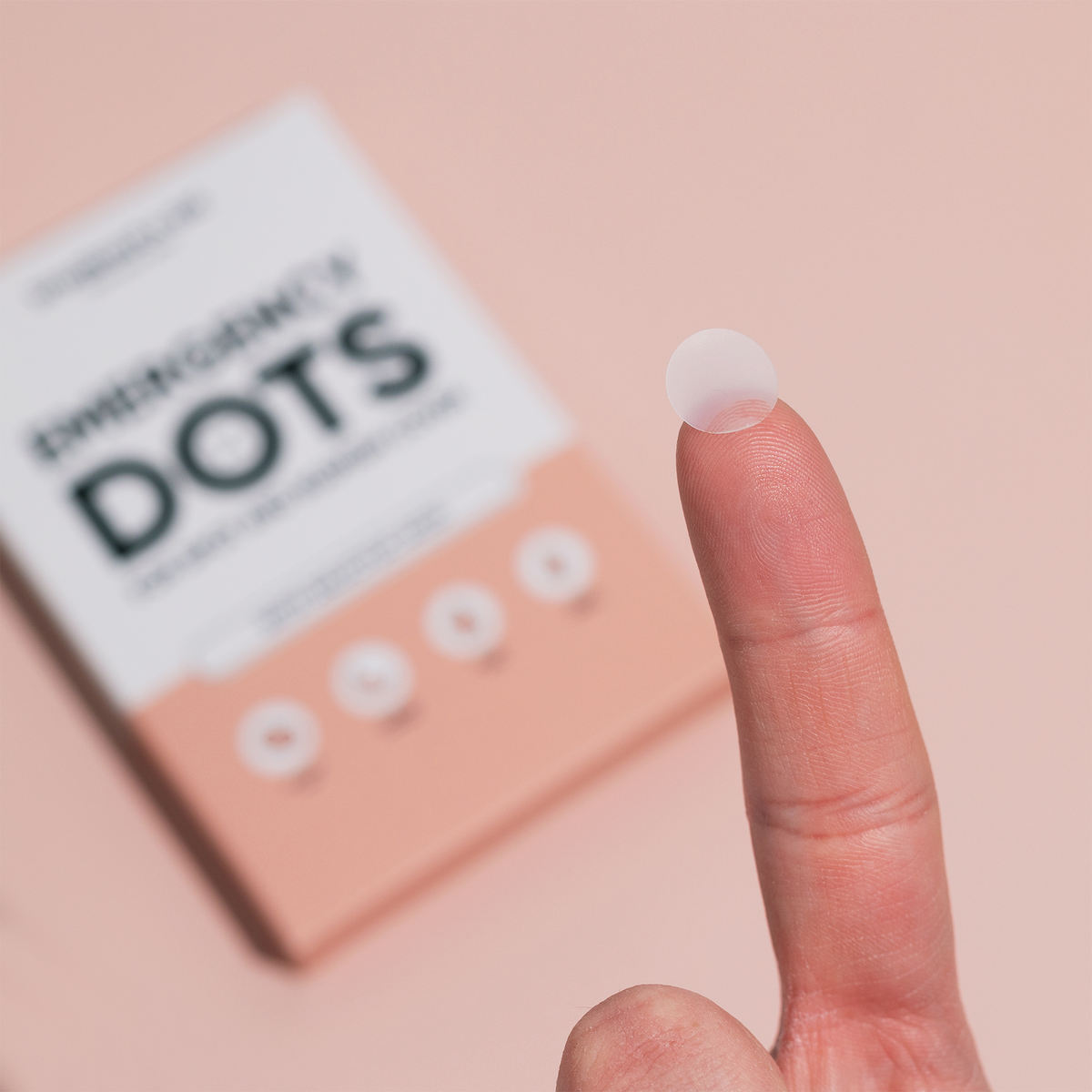 Ein kleines, durchscheinendes Emergency Dots with Salicylic Acid-Pflaster auf der Fingerspitze, im Hintergrund die Verpackung.